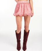 A model is shown from the waist down wearing a pink and red floral print bubble short paired with dark burgundy suede knee-high boots, against a plain white studio background.