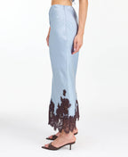 A woman modeling a light blue tank and midi skirt  with brown floral embroidery and appliqué on the bodice and hem, set against a plain white studio background.
