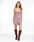 A woman modeling a short, sleeveless dusty rose snakeskin print dress with brown knee-high boots, standing against a plain white studio background.