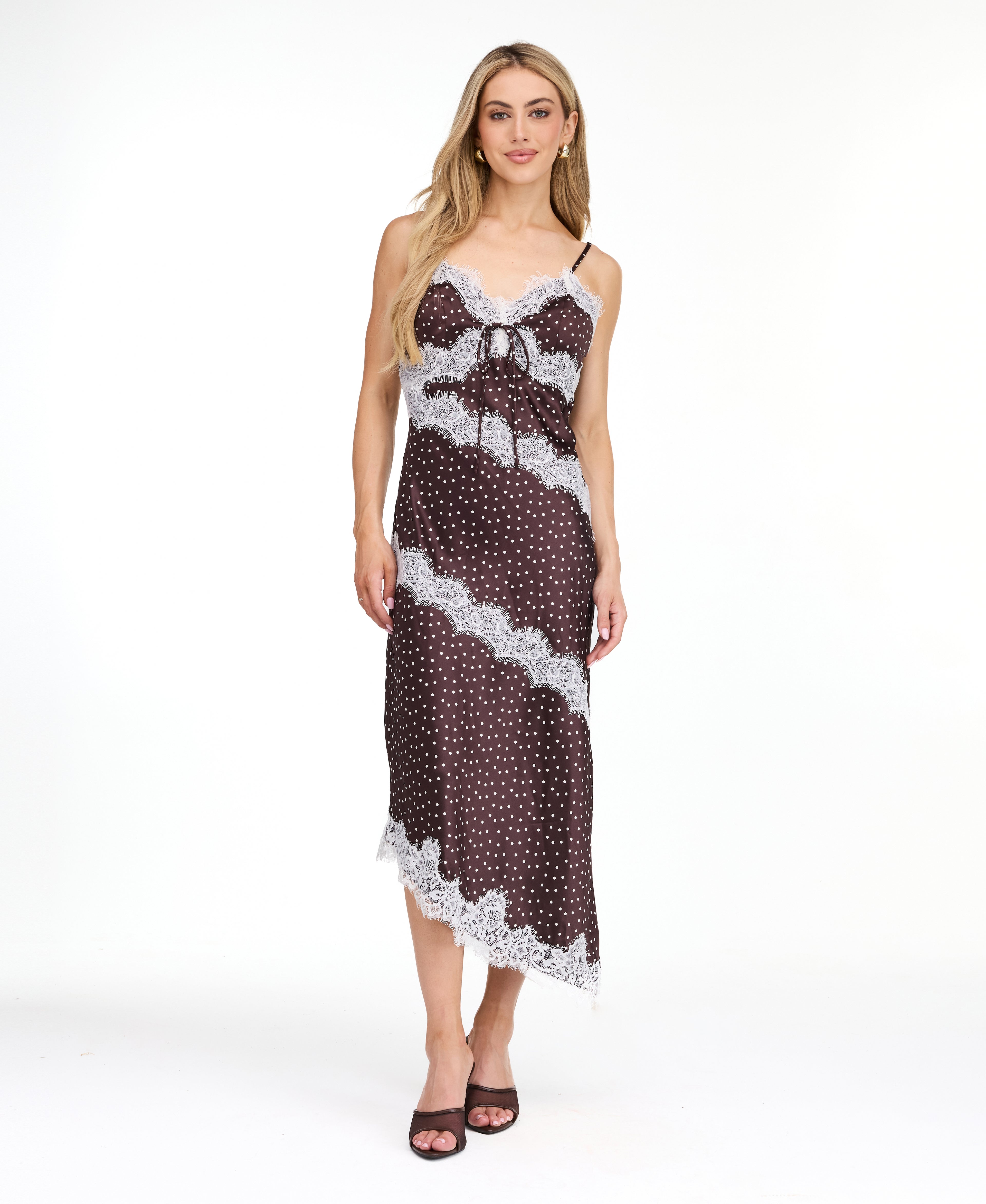 A woman modeling a dark brown and white polka dot slip dress with white lace trim, photographed against a solid white studio background.