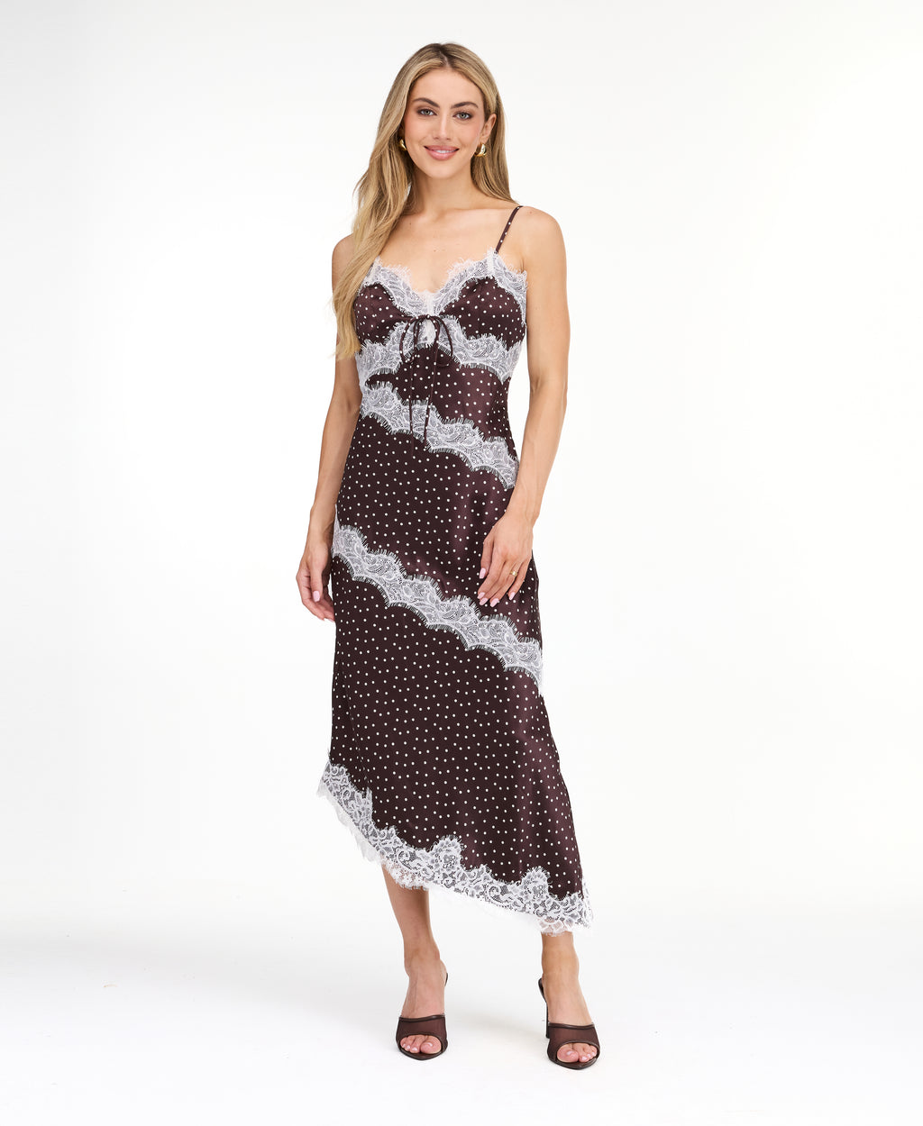 A woman modeling a dark brown and white polka dot slip dress with white lace trim, photographed against a solid white studio background.