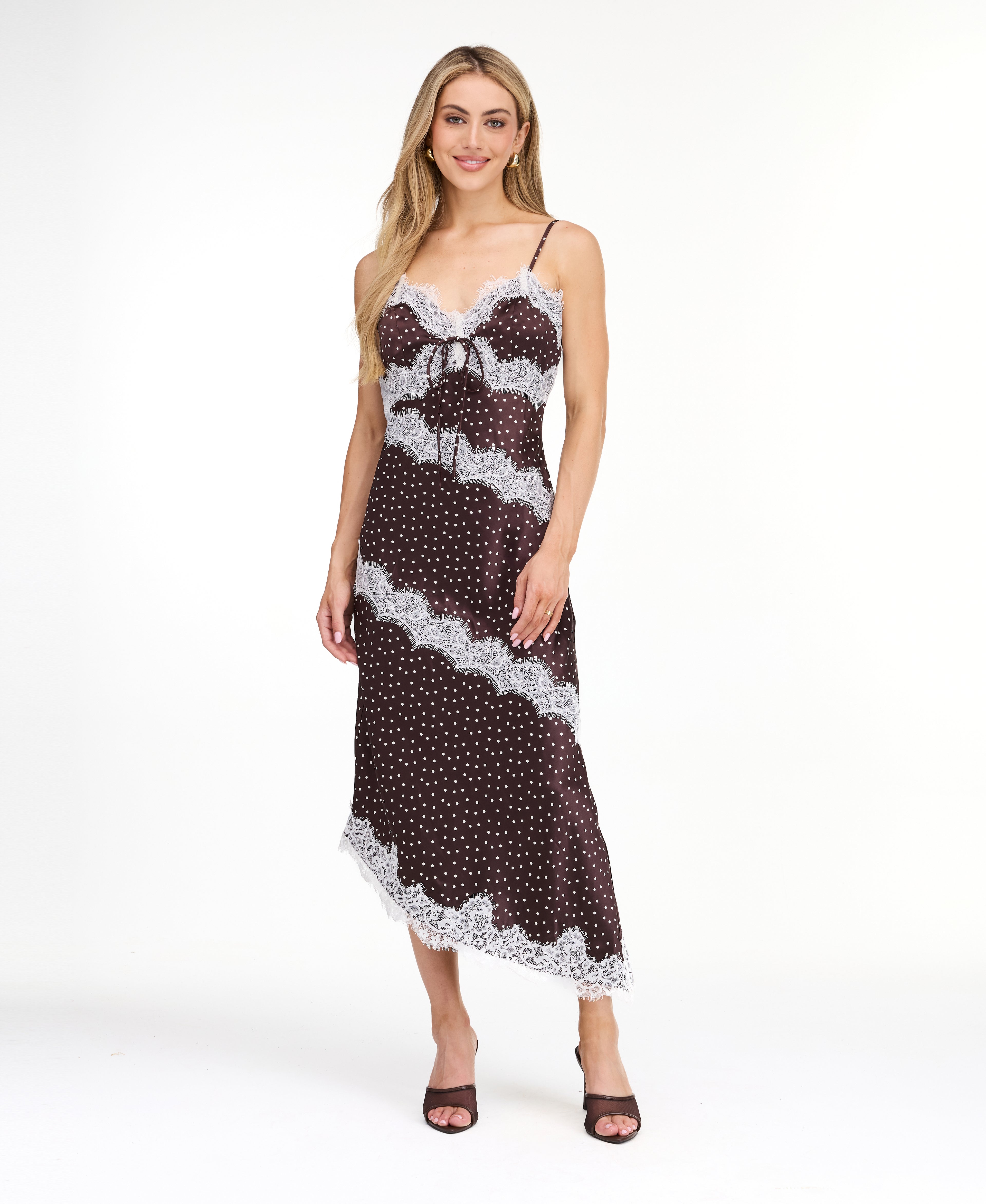 A woman modeling a dark brown and white polka dot slip dress with white lace trim, photographed against a solid white studio background.