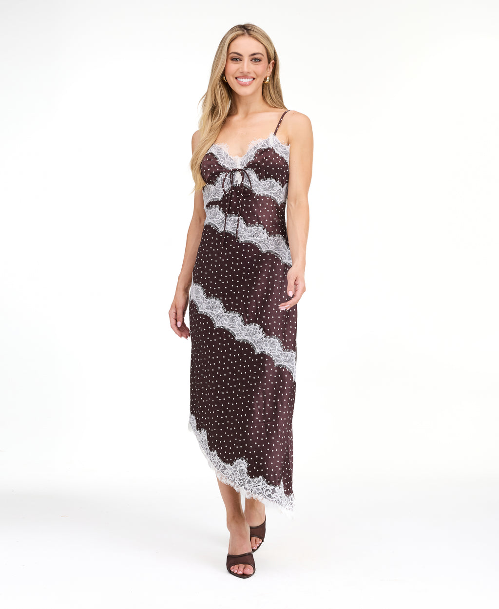 A woman modeling a dark brown and white polka dot slip dress with white lace trim, photographed against a solid white studio background.