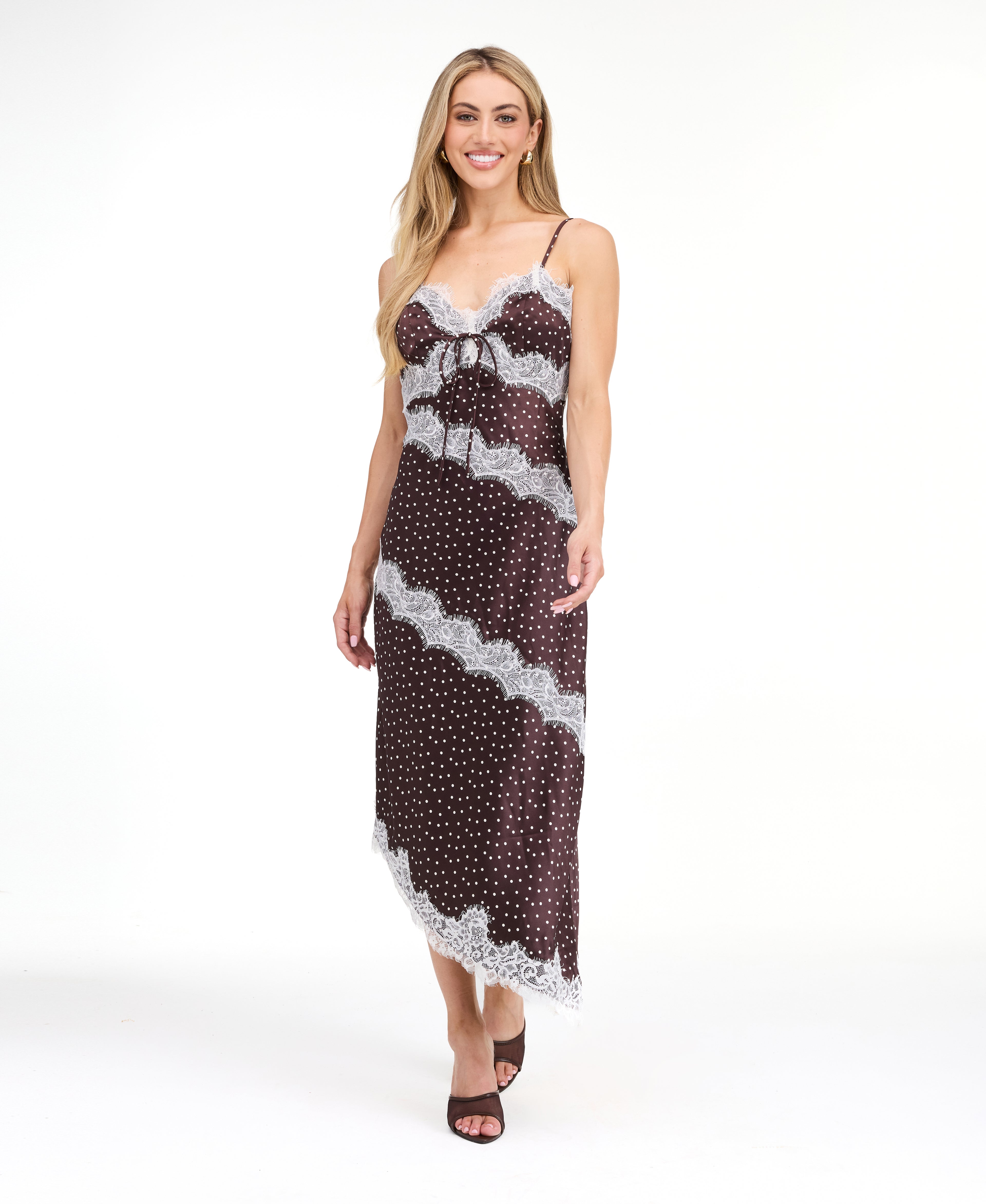 A woman modeling a dark brown and white polka dot slip dress with white lace trim, photographed against a solid white studio background.