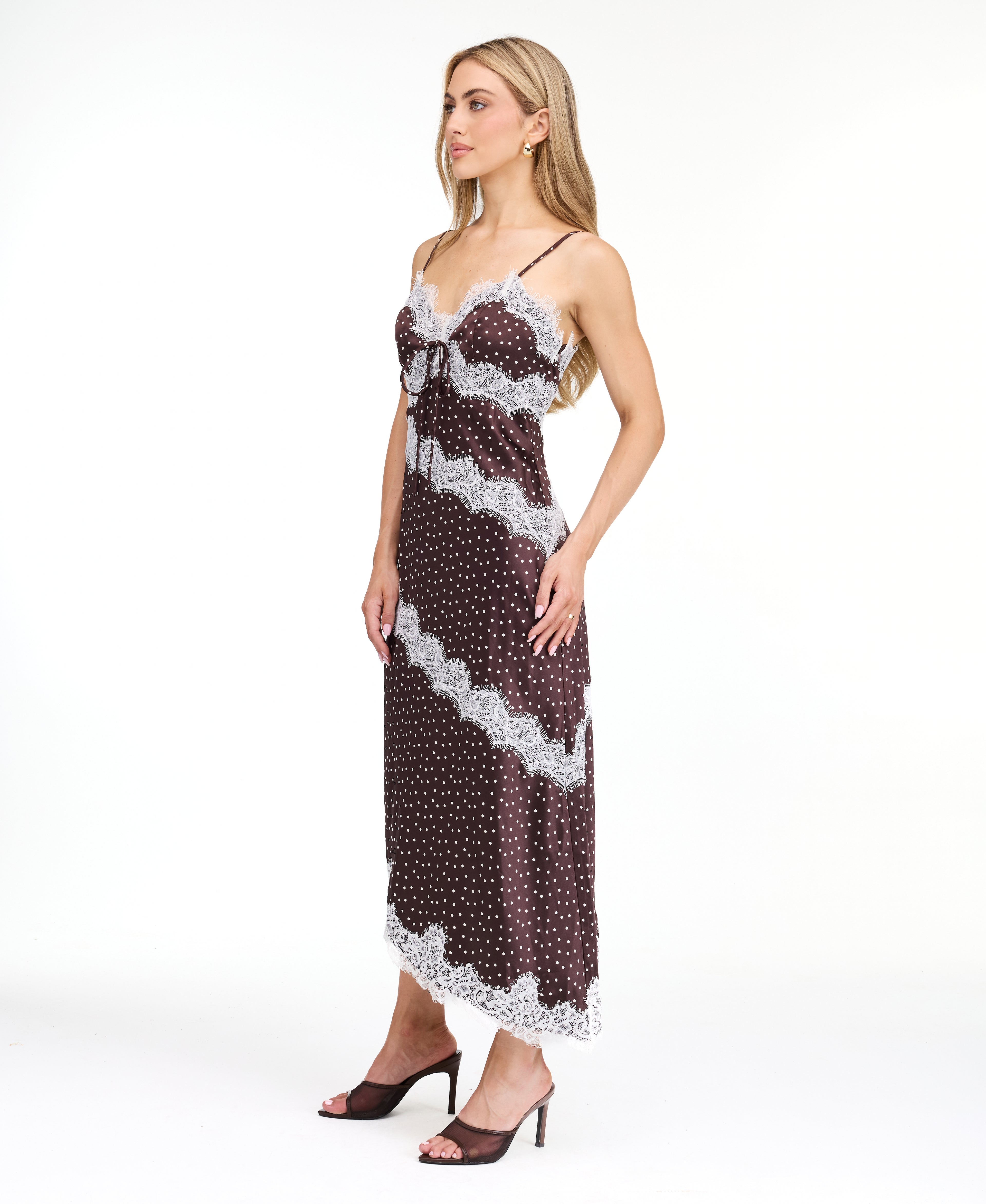 A woman modeling a dark brown and white polka dot slip dress with white lace trim, photographed against a solid white studio background.