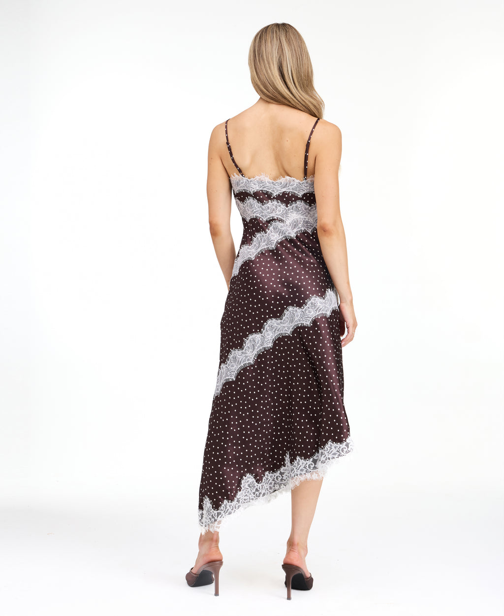 A woman modeling a dark brown and white polka dot slip dress with white lace trim, photographed against a solid white studio background.