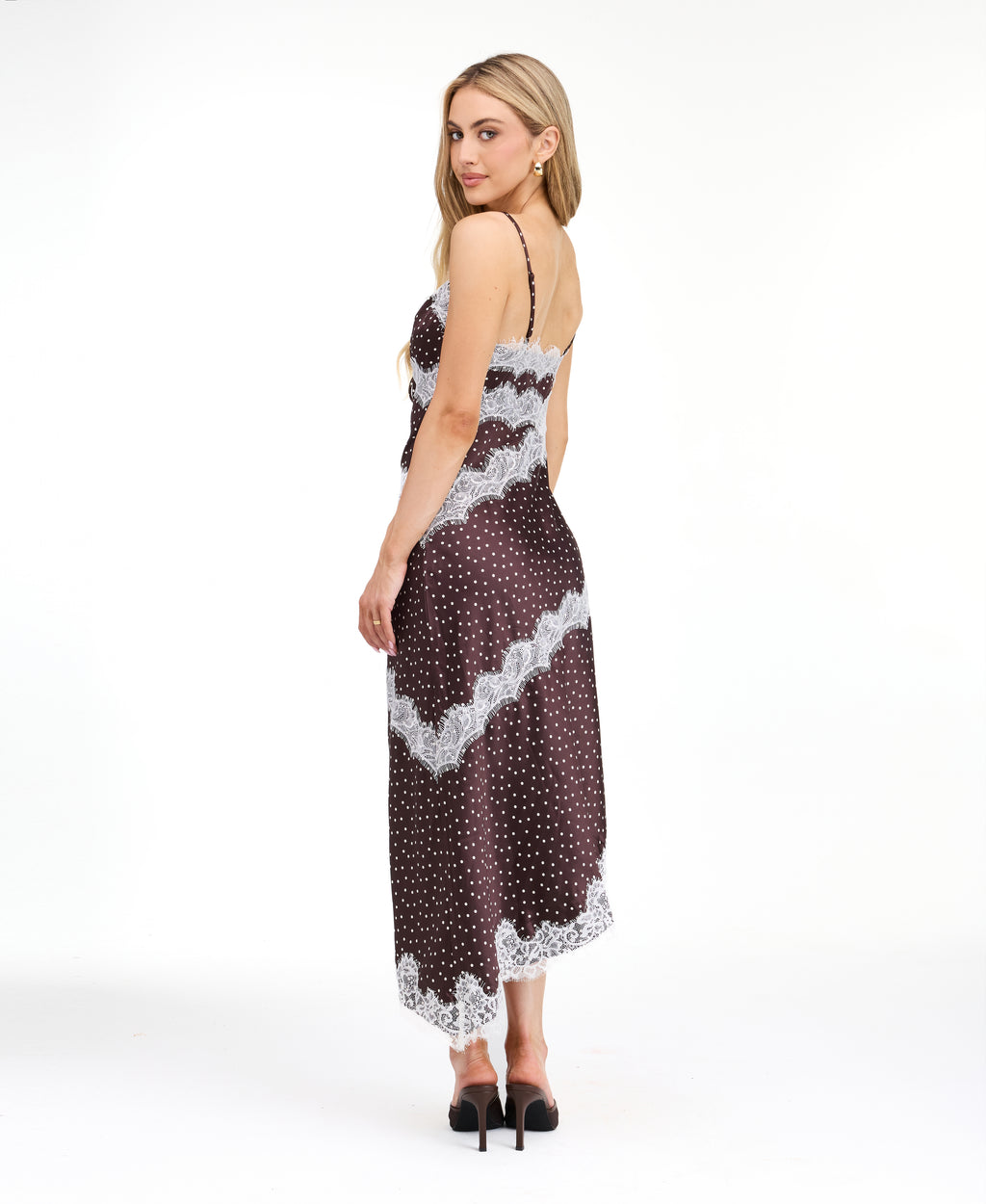 A woman modeling a dark brown and white polka dot slip dress with white lace trim, photographed against a solid white studio background.