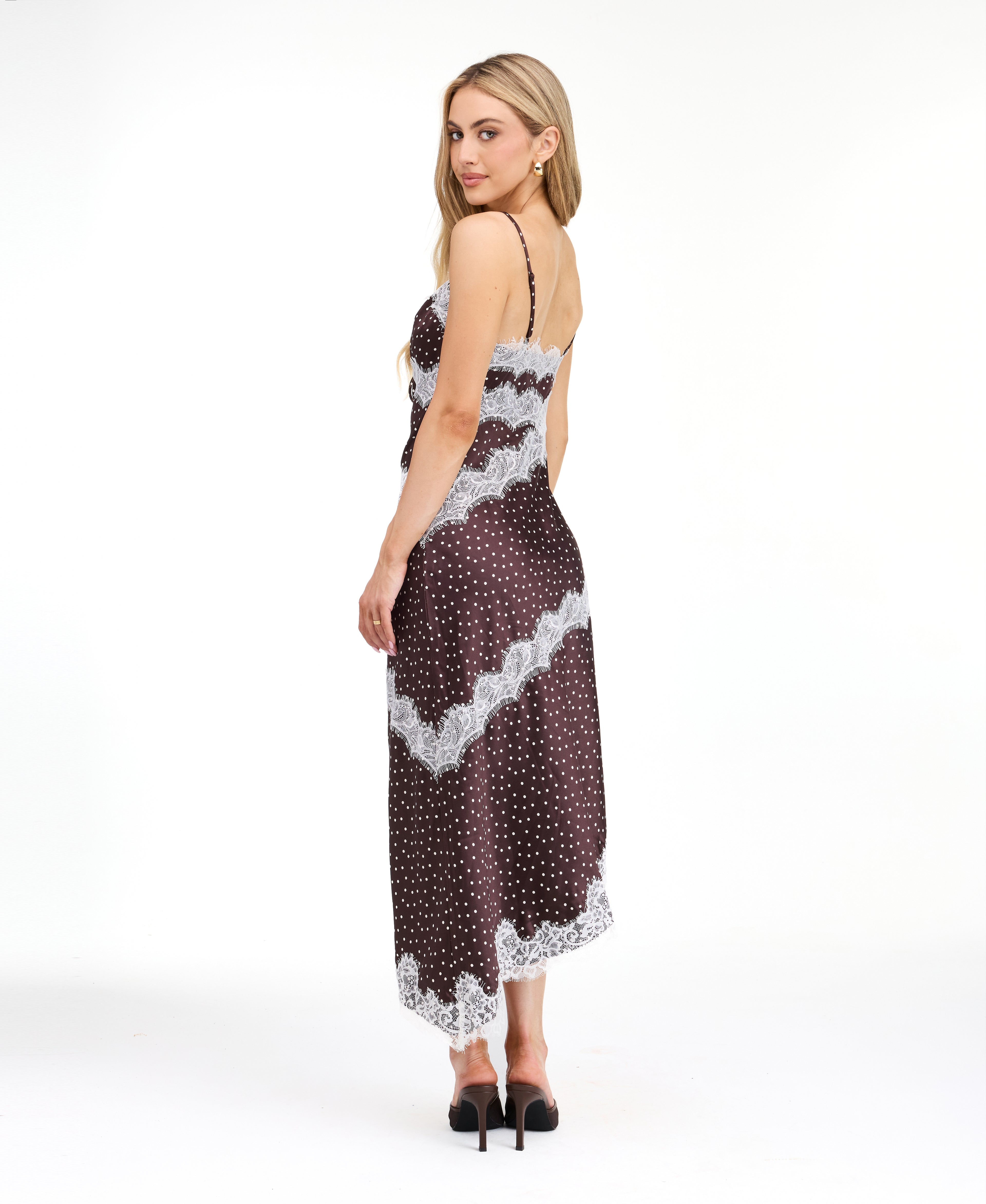 A woman modeling a dark brown and white polka dot slip dress with white lace trim, photographed against a solid white studio background.