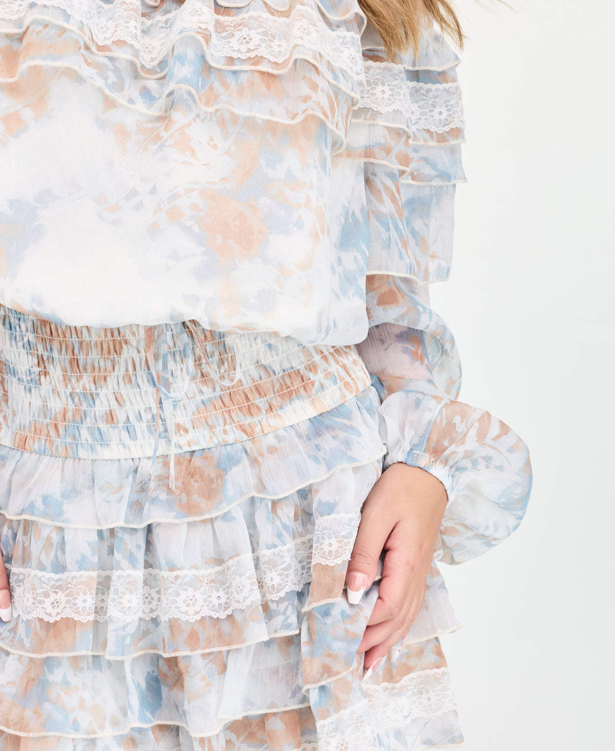 A woman modeling an off-the-shoulder tiered mini dress with a light blue, white, and beige abstract print, paired with brown suede knee-high boots, against a plain white studio background.