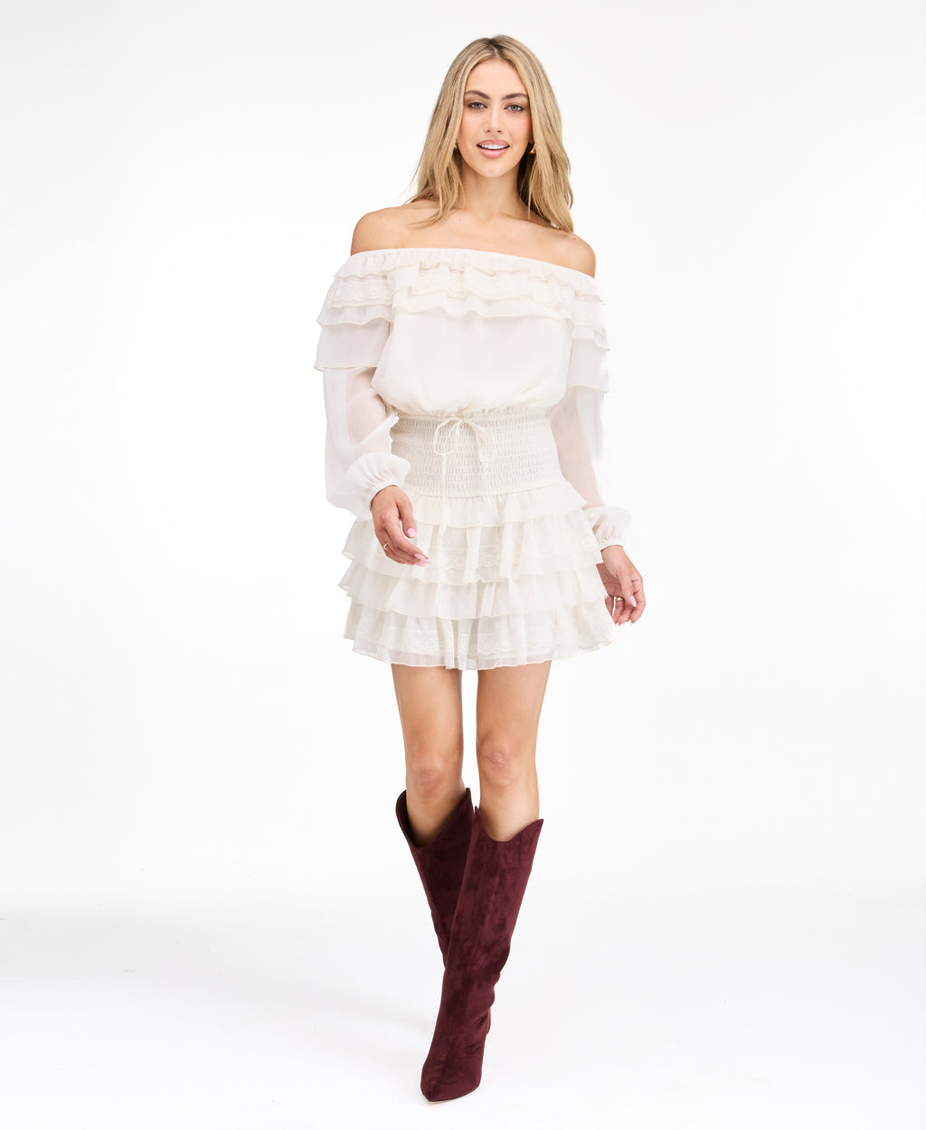 A model wearing an ivory off shoulder dress with shear bubble sleeves. The dress consists of a  smocked waistband and ruffle lace details throughout the neckline and skirt 