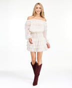 A model wearing an ivory off shoulder dress with shear bubble sleeves. The dress consists of a  smocked waistband and ruffle lace details throughout the neckline and skirt 