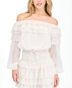 A model wearing an ivory off shoulder dress with shear bubble sleeves. The dress consists of a  smocked waistband and ruffle lace details throughout the neckline and skirt 