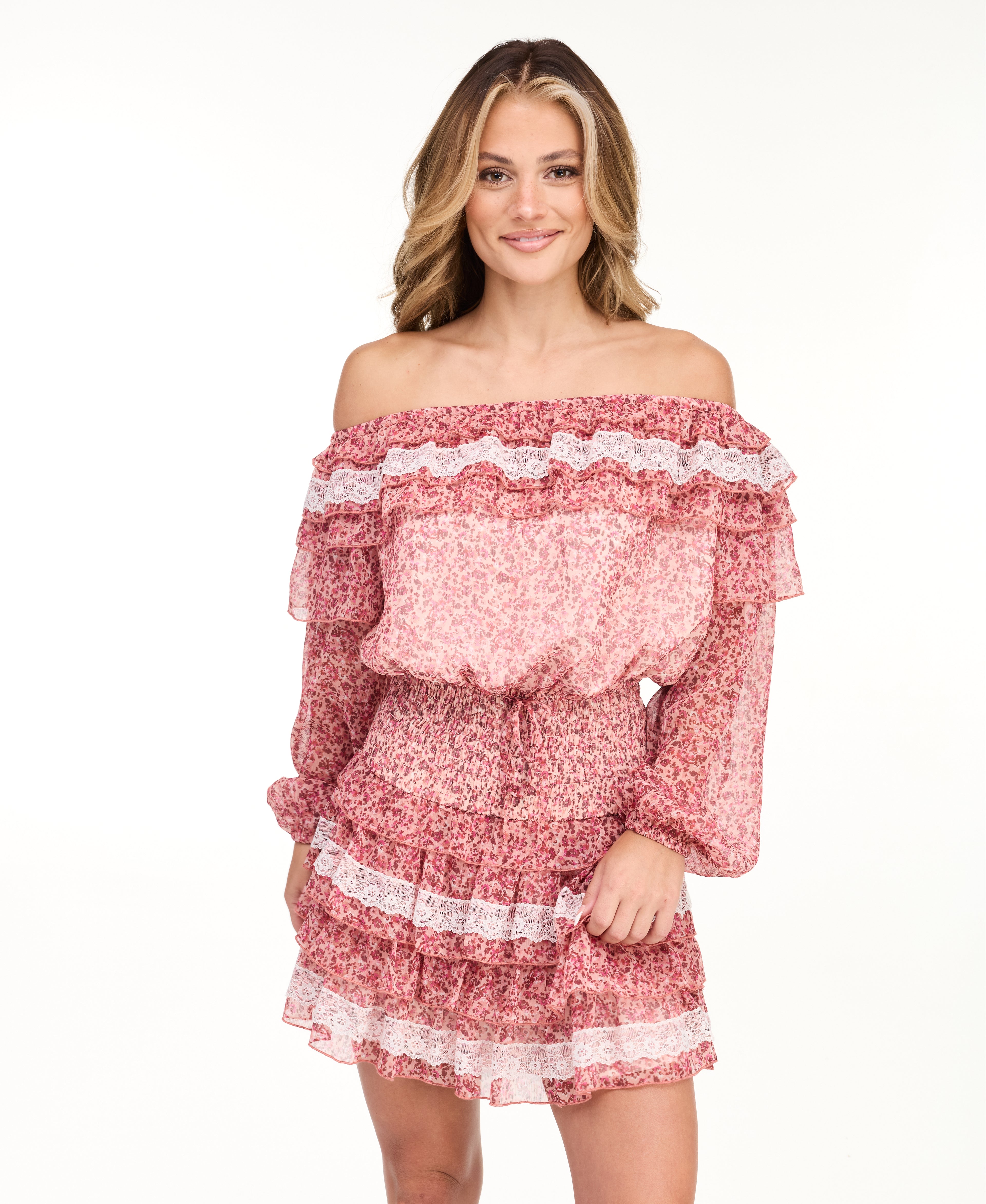 A woman modeling an off-the-shoulder long-sleeve mini dress with a pink and white floral pattern, paired with dark brown knee-high boots, against a plain white studio background.