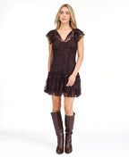 A woman modeling a dark brown lace mini dress with ruffled cap sleeves and tie detail at the neckline, paired with dark brown knee-high boots, against a plain white studio background.