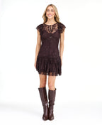 A woman modeling a dark brown lace mini dress with ruffled cap sleeves and tie detail at the neckline, paired with dark brown knee-high boots, against a plain white studio background.