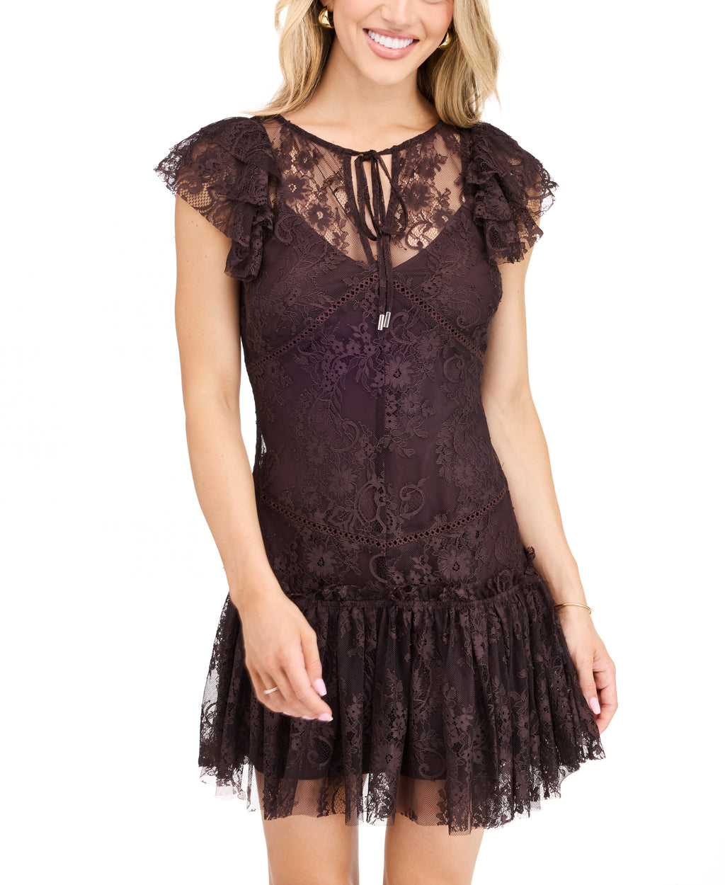 A woman modeling a dark brown lace mini dress with ruffled cap sleeves and tie detail at the neckline, paired with dark brown knee-high boots, against a plain white studio background.