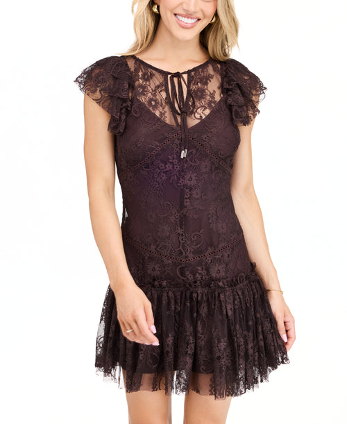 A woman modeling a dark brown lace mini dress with ruffled cap sleeves and tie detail at the neckline, paired with dark brown knee-high boots, against a plain white studio background.