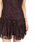 A woman modeling a dark brown lace mini dress with ruffled cap sleeves and tie detail at the neckline, paired with dark brown knee-high boots, against a plain white studio background.