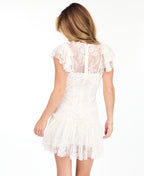 A woman modeling a short white lace dress with ruffled cap sleeves and brown knee-high suede boots, standing against a plain white studio background.