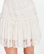 A woman modeling a short white lace dress with ruffled cap sleeves and brown knee-high suede boots, standing against a plain white studio background.