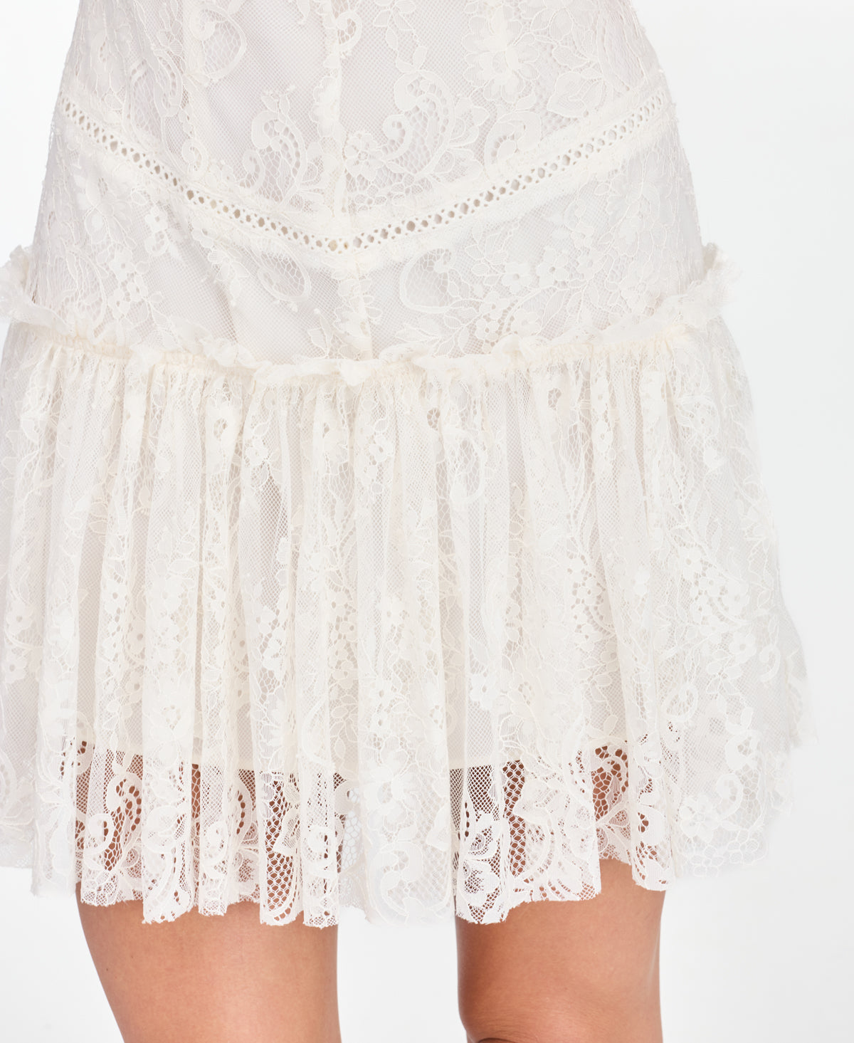A woman modeling a short white lace dress with ruffled cap sleeves and brown knee-high suede boots, standing against a plain white studio background.