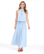 A woman modeling a light blue sleeveless maxi dress with a smocked waist and with a halter neck scarf, set against a plain white studio background.
