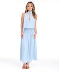 A woman modeling a light blue sleeveless maxi dress with a smocked waist and with a halter neck scarf, set against a plain white studio background.