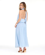 A woman modeling a light blue sleeveless maxi dress with a smocked waist and with a halter neck scarf, set against a plain white studio background.