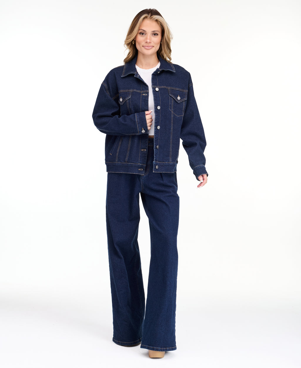 A woman modeling a dark denim wash jacket and matching wide-leg jeans over a white top, photographed against a seamless white studio background.