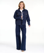 A woman modeling a dark denim wash jacket and matching wide-leg jeans over a white top, photographed against a seamless white studio background.
