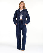 A woman modeling a dark denim wash jacket and matching wide-leg jeans over a white top, photographed against a seamless white studio background.