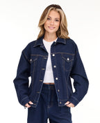 A woman modeling a dark denim wash jacket and matching wide-leg jeans over a white top, photographed against a seamless white studio background.
