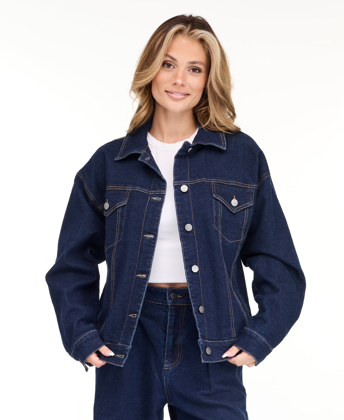A woman modeling a dark denim wash jacket and matching wide-leg jeans over a white top, photographed against a seamless white studio background.