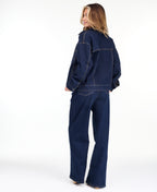 A woman modeling a dark denim wash jacket and matching wide-leg jeans over a white top, photographed against a seamless white studio background.