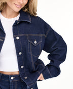 A woman modeling a dark denim wash jacket and matching wide-leg jeans over a white top, photographed against a seamless white studio background.