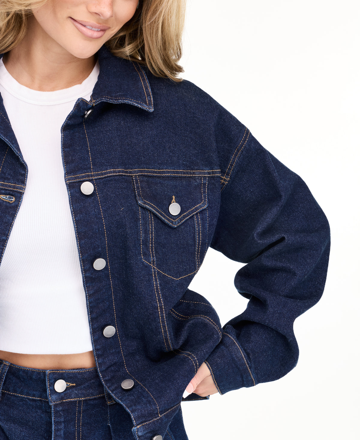 A woman modeling a dark denim wash jacket and matching wide-leg jeans over a white top, photographed against a seamless white studio background.