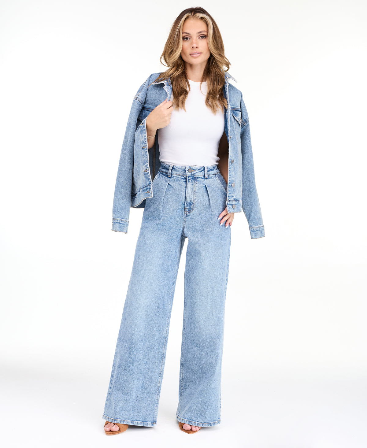 A woman modeling a light-wash blue denim outfit consisting of wide-leg jeans, a white ribbed top, and an open light blue denim jacket, photographed against a seamless white studio background.