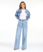 A woman modeling a light-wash blue denim outfit consisting of wide-leg jeans, a white ribbed top, and an open light blue denim jacket, photographed against a seamless white studio background.