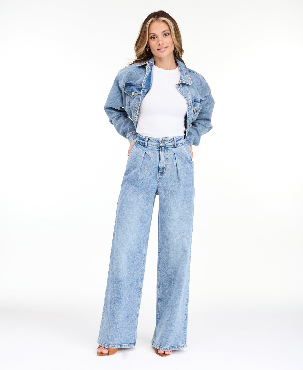 A woman modeling a light-wash blue denim outfit consisting of wide-leg jeans, a white ribbed top, and an open light blue denim jacket, photographed against a seamless white studio background.