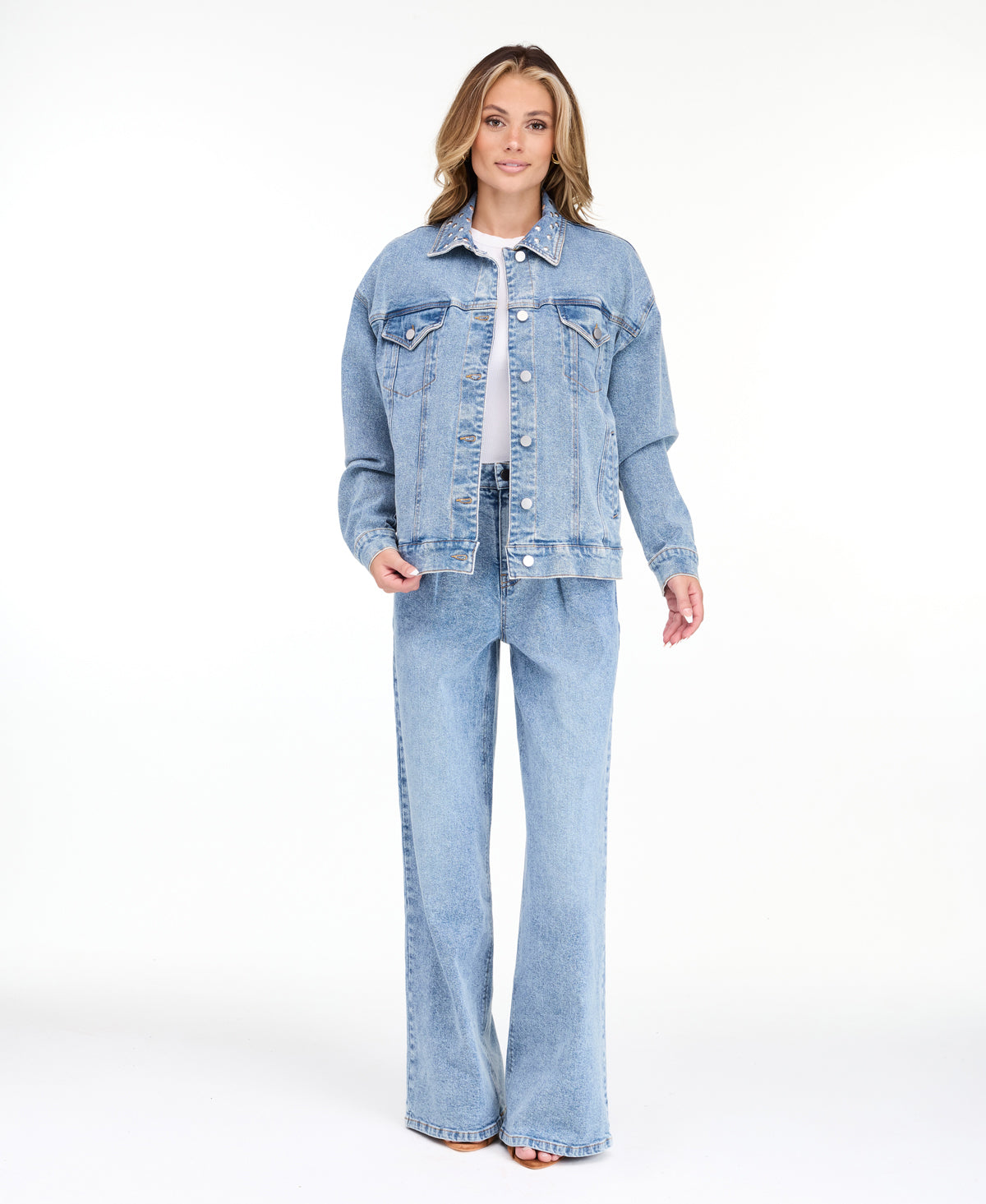 A woman modeling a light blue denim jacket with studs on the neckline and matching wide-leg jeans against a plain white studio background.
