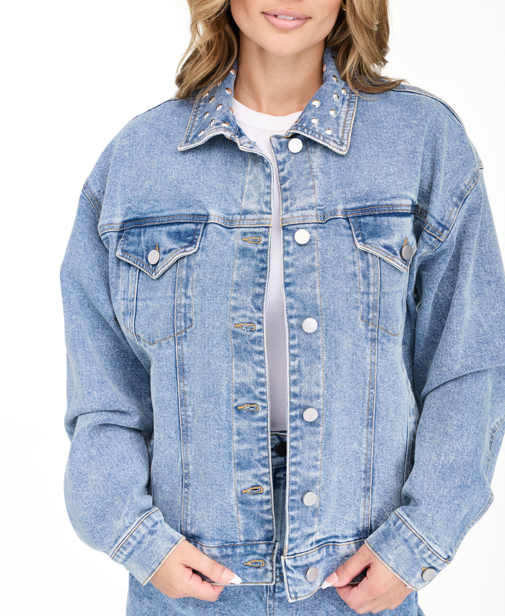 A woman modeling a light blue denim jacket with studs on the neckline and matching wide-leg jeans against a plain white studio background.