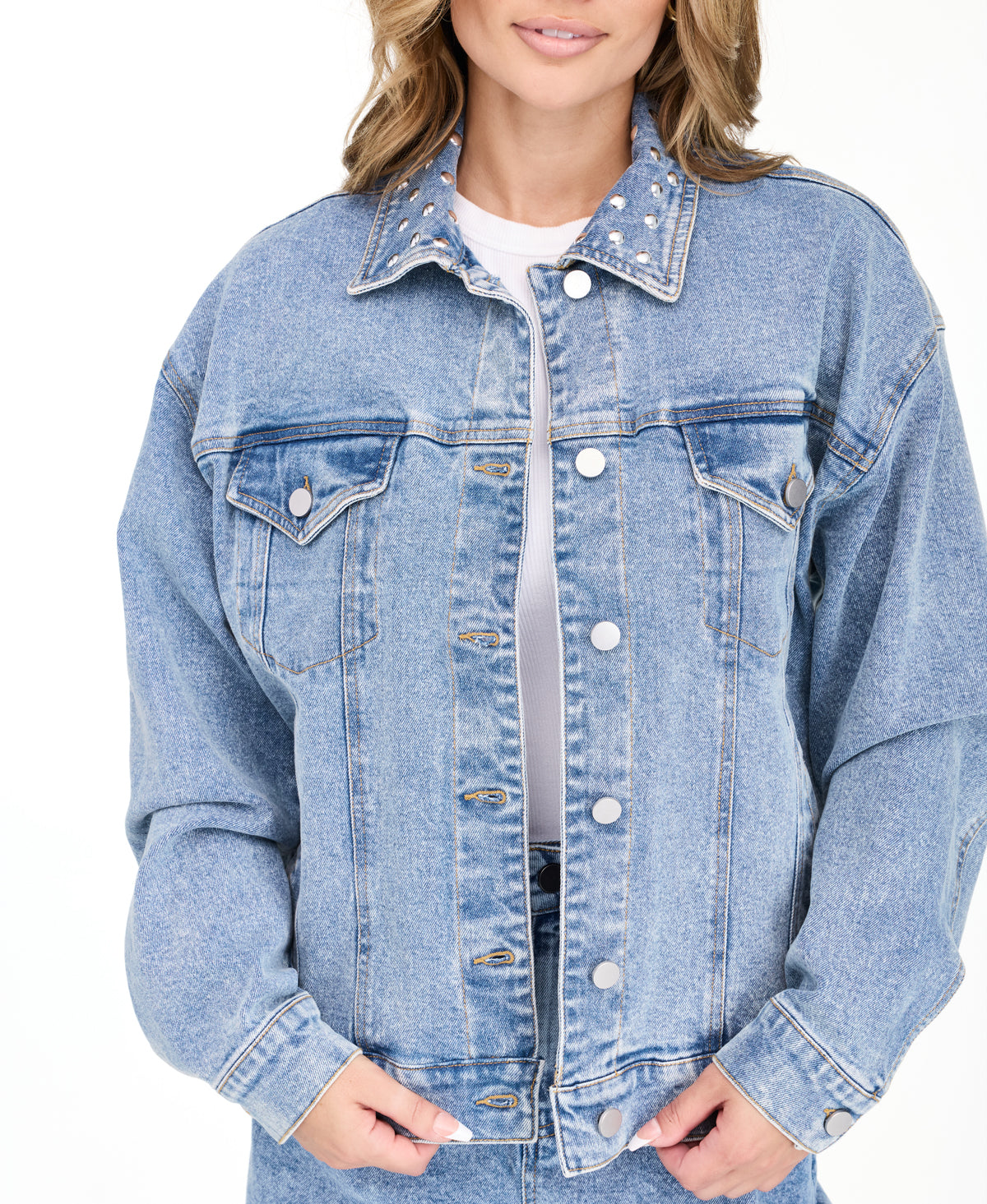 A woman modeling a light blue denim jacket with studs on the neckline and matching wide-leg jeans against a plain white studio background.