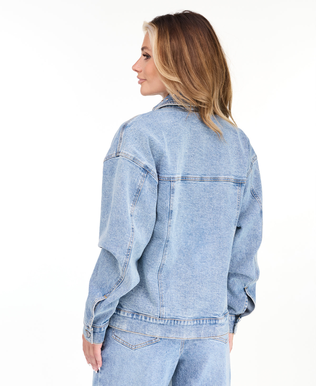 A woman modeling a light blue denim jacket with studs on the neckline and matching wide-leg jeans against a plain white studio background.