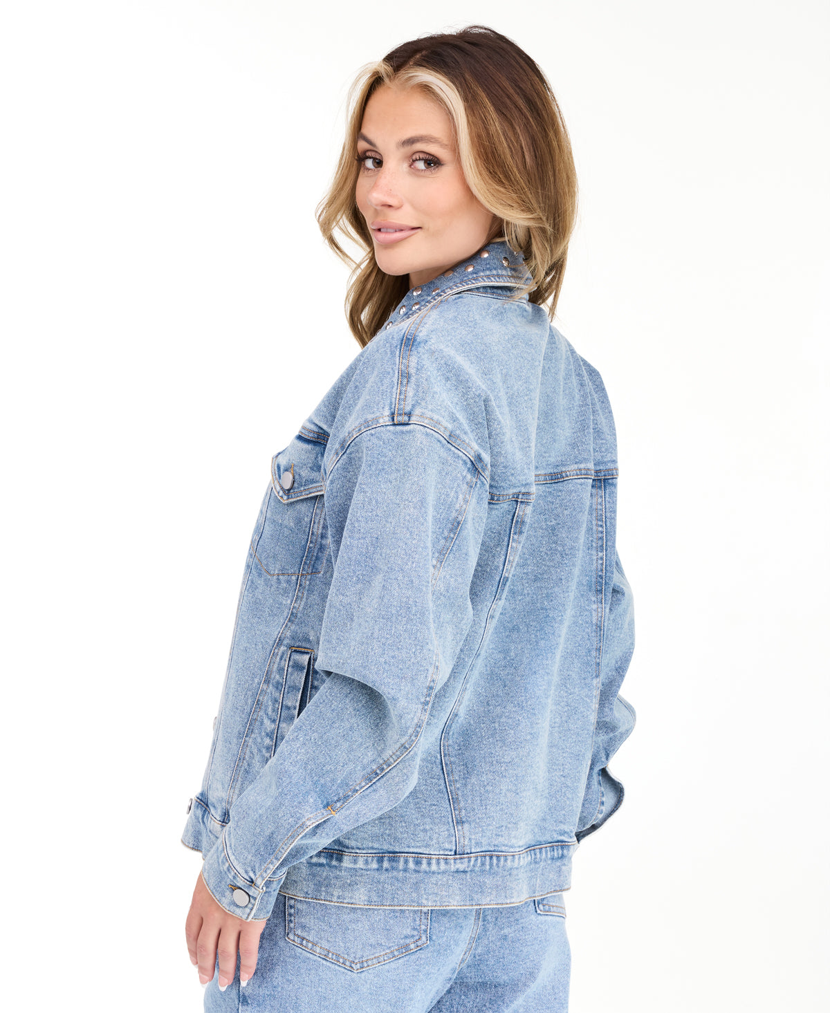 A woman modeling a light blue denim jacket with studs on the neckline and matching wide-leg jeans against a plain white studio background.