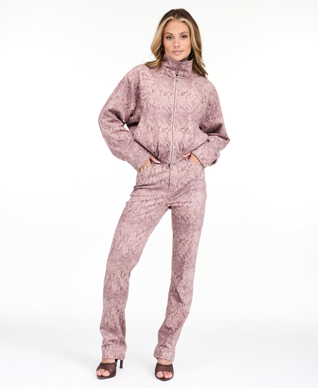 A woman modeling a dusty rose pink and brown snakeskin pattern, featuring a cropped zip-up jacket and matching pants, set against a plain white studio background.