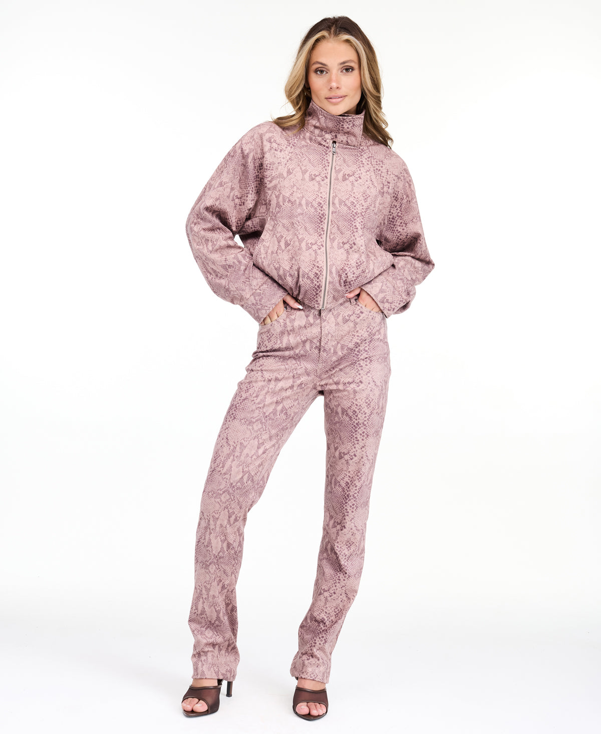 A woman modeling a dusty rose pink and brown snakeskin pattern, featuring a cropped zip-up jacket and matching pants, set against a plain white studio background.