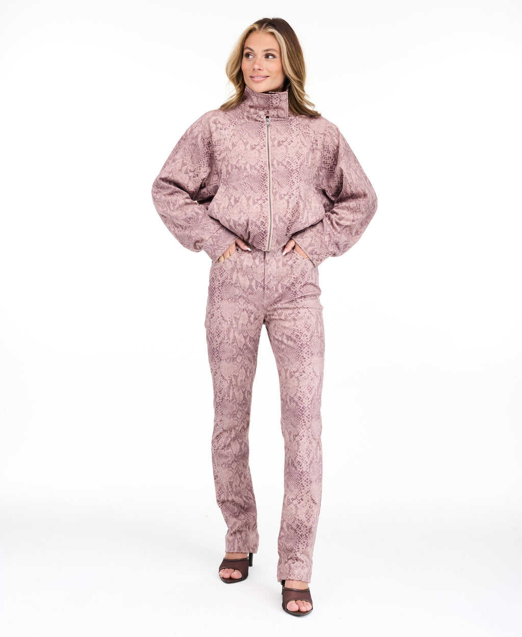 A woman modeling a dusty rose pink and brown snakeskin pattern, featuring a cropped zip-up jacket and matching pants, set against a plain white studio background.