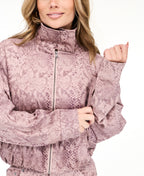 A woman wearing a dusty rose snakeskin print zip-up jacket with a mock neck, paired with pants, against a plain white background.
