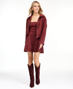 A woman modeling a matching burgundy zip-up jacket, fit and flare dress,  and tall knee-high boots against a plain white studio background.
