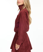 A woman modeling a matching burgundy zip-up jacket, fit and flare dress,  and tall knee-high boots against a plain white studio background.